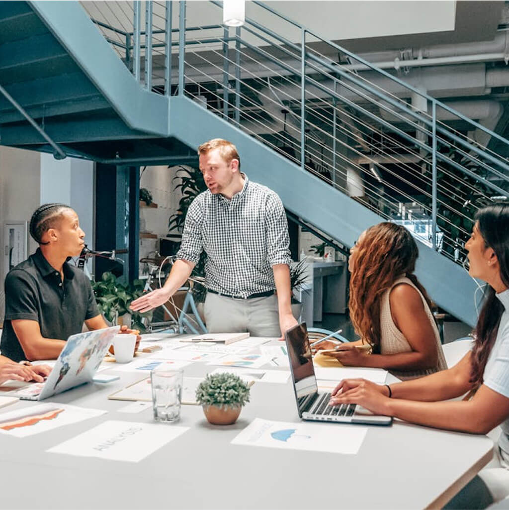 Four people sit at a table with laptops and documents, while one person—possibly a Ph.D.—stands and speaks to them in a modern office setting under a staircase.