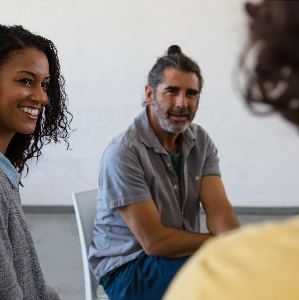 Three people sit in a circle indoors, engaging in conversation. The woman smiles warmly, while the man in the middle listens intently—an inviting scene that captures the spirit of Art Therapy.