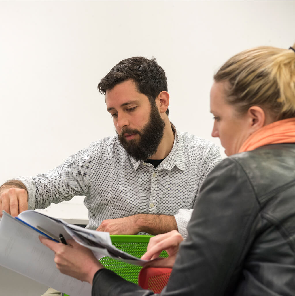 A man and a woman review documents together at a desk; the man gestures toward a page while the woman, possibly discussing Art Therapy, holds the papers.