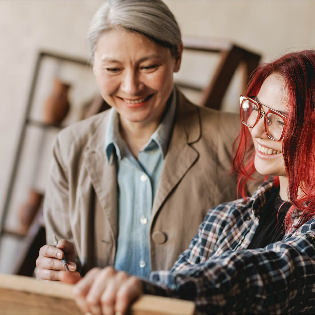 Two women smiling and looking at a wooden board together indoors—one with Grey hair in a blazer, the other with red hair and glasses in a flannel shirt—explore creative ideas as part of an Art Therapy PhD session.