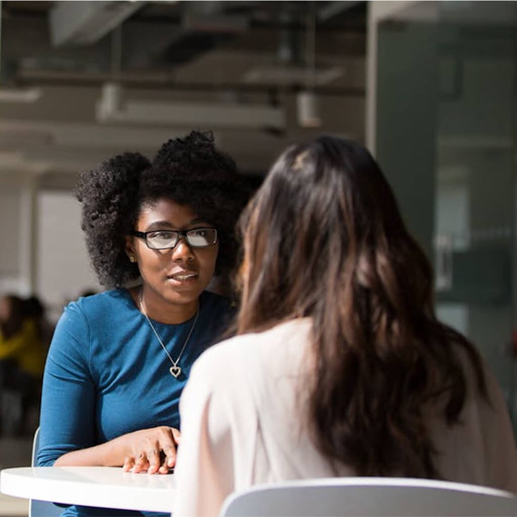 Two women sit across from each other at a table in an office setting, engaged in conversation about Art Therapy.