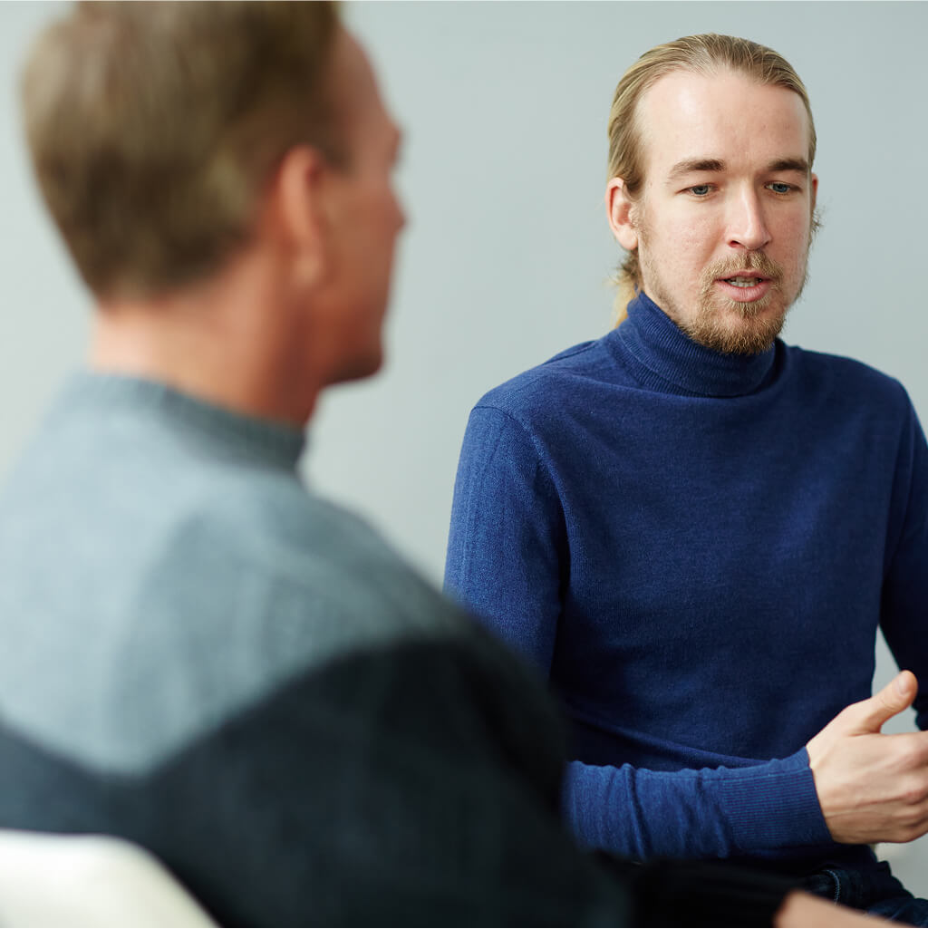 Two men are sitting indoors having a serious conversation; one, possibly a Ph.D. professional, faces the camera while the other is turned away and slightly blurred.