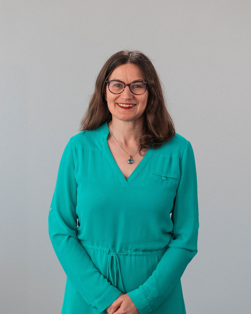 Nicole Dorfan, una mujer de pelo castaño largo, gafas y vestido verde azulado, está de pie sobre un fondo claro y liso, sonriendo con las manos cruzadas al frente.