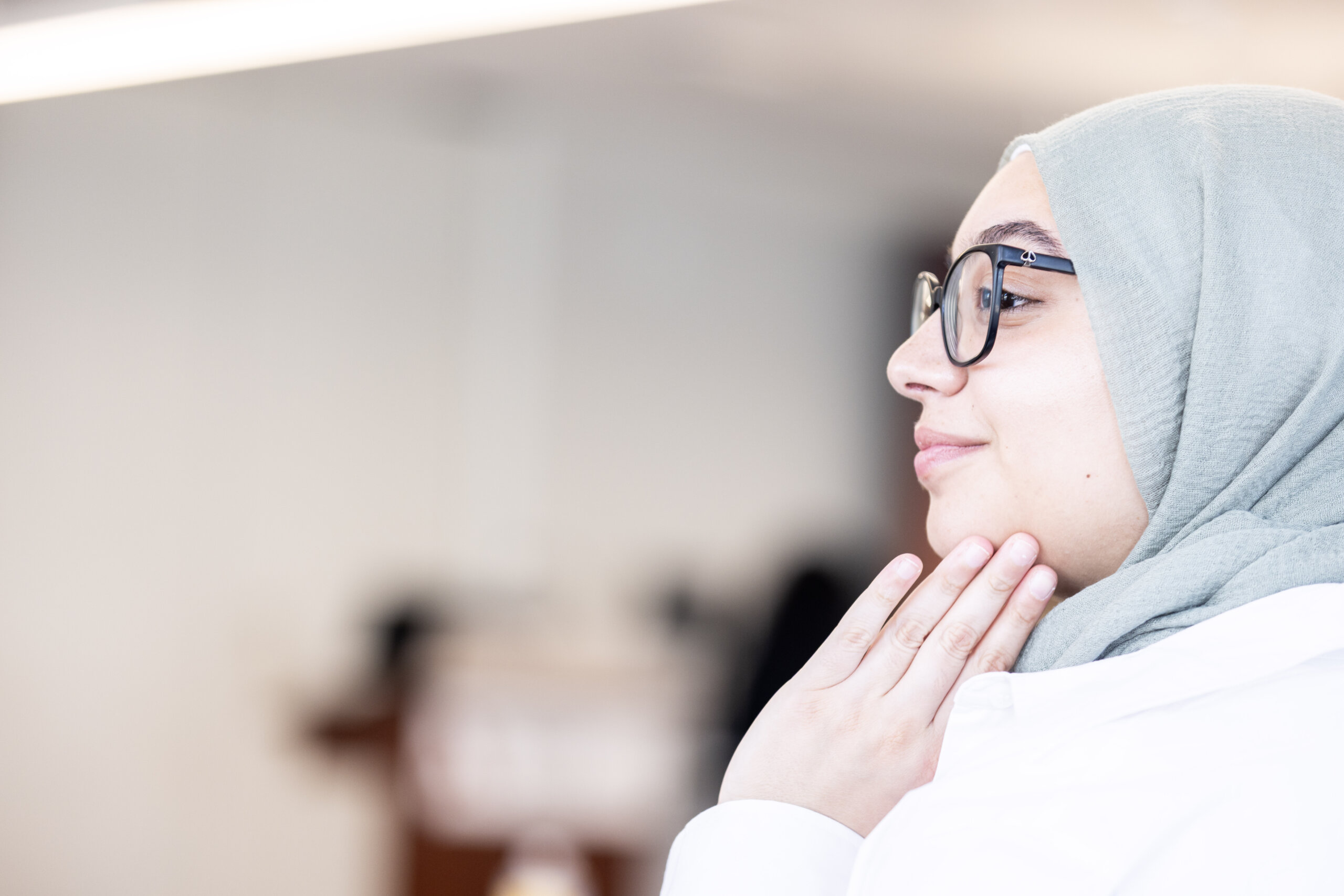 A woman wearing glasses and a light green hijab—perhaps a Clinical Mental Health Counselling, M.A. student—touches her neck and looks to the side, set against a blurred indoor background.