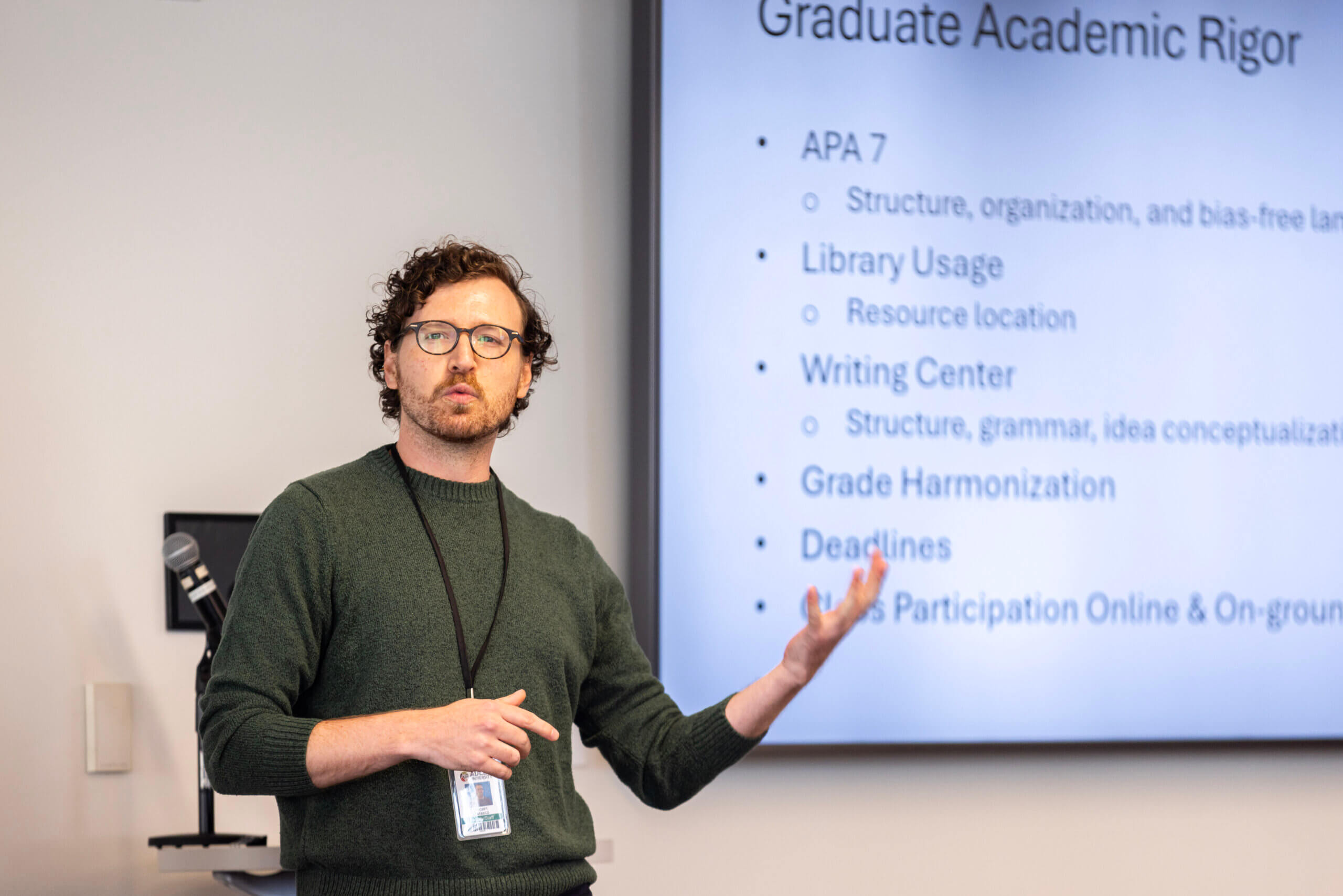 A man gives a presentation in a classroom, gesturing toward a projected slide titled “Graduate Academic Rigor” with bullet points about APA 7, library usage, and deadlines for the M.A. in Clinical Mental Health Counseling program.