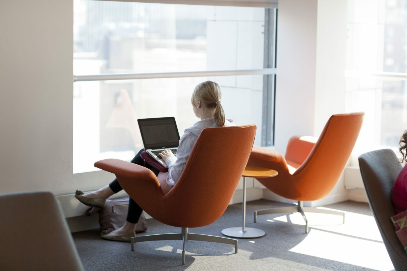 A person sits in an orange chair by a large window, working on a laptop in a modern, well-lit room.