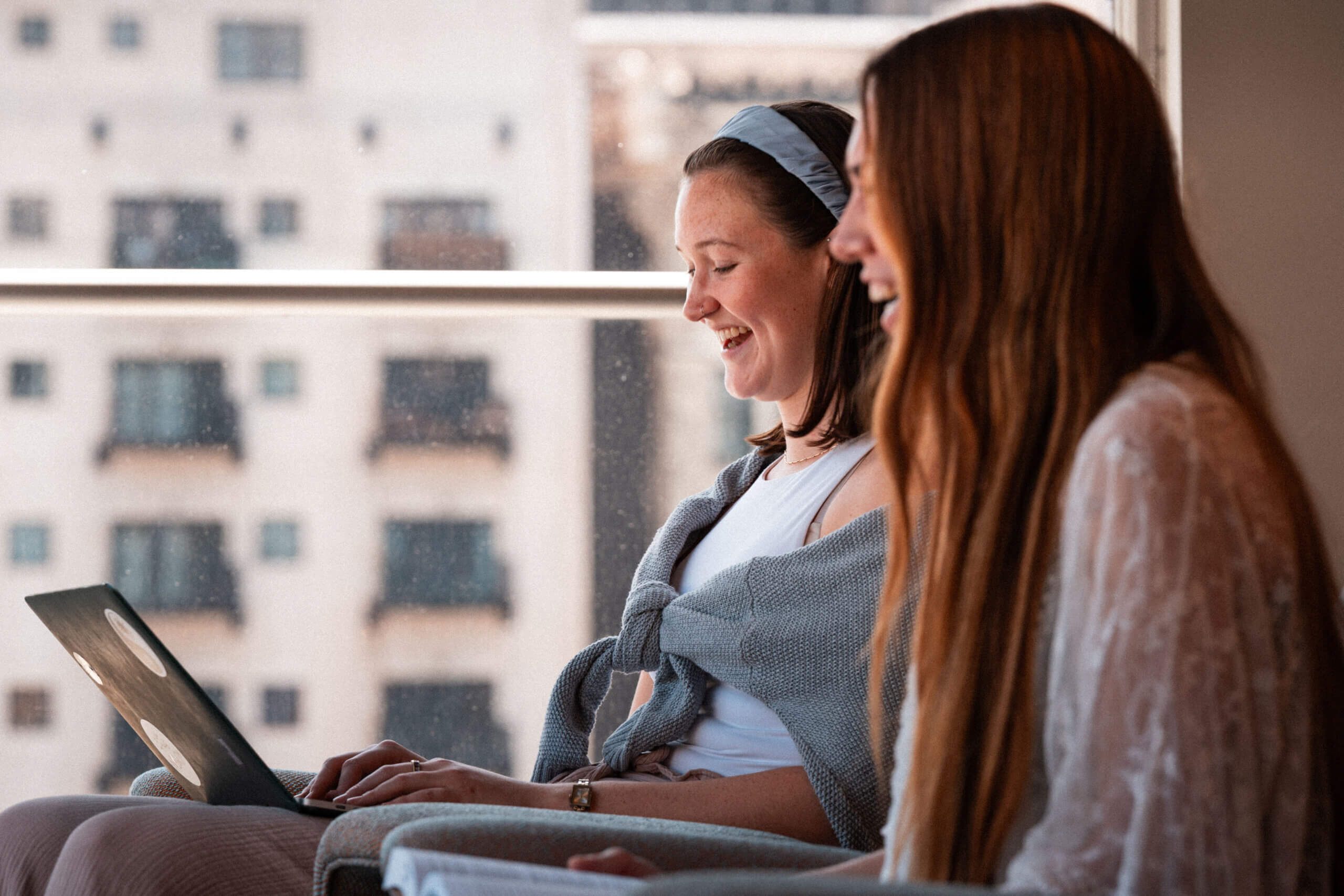 Two women sit side by side near a window, smiling and looking at a laptop. An urban building is visible in the background through the glass. The scene appears relaxed and cheerful.