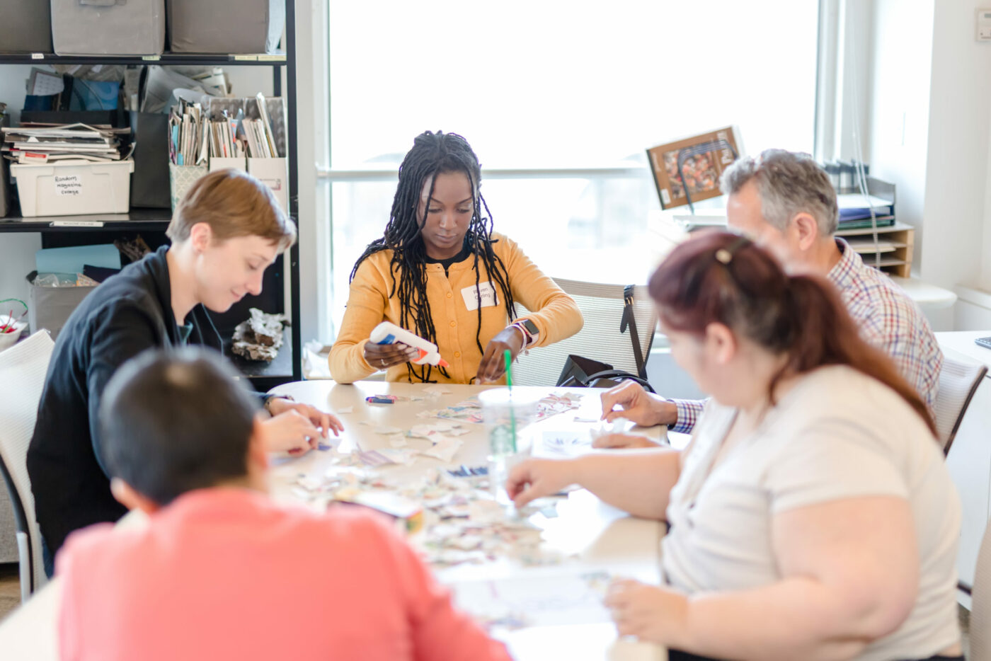 A group of five people sit around a table, working on an arts and crafts project with paper and scissors in a bright, organized room that reflects contemporary Practise inspired by Adler’s principles.