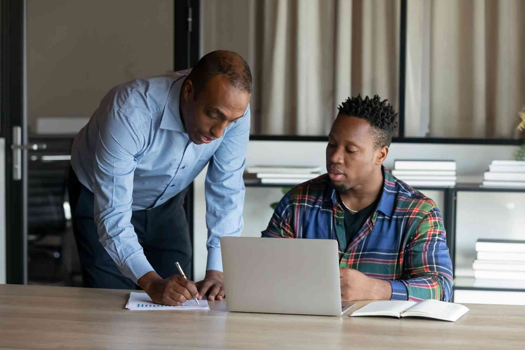 Two men work together at a desk; one is sitting with a laptop and notebook, while the other stands, writing on paper and looking at the computer screen.