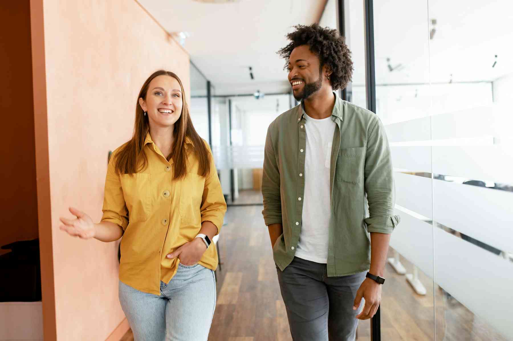 Two people walk and talk in a modern office hallway. The woman gestures with her hand while the man listens and smiles. Both appear relaxed and engaged in conversation.
