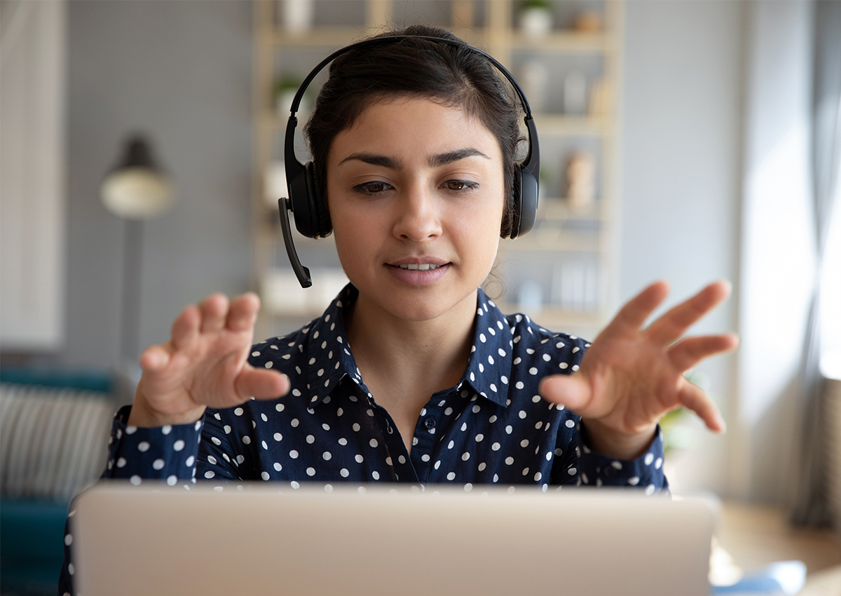 Mujer con auriculares sentada frente a un ordenador portátil, gesticulando con las manos mientras participa en una videollamada en un entorno de oficina en casa, adaptándose durante una crisis con confianza y comunicación eficaz.