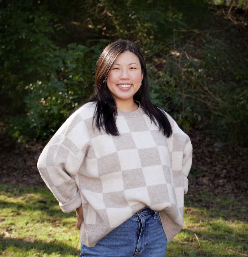 A woman stands outdoors on grass, smiling, wearing a beige and white checkered sweater and blue jeans. An Adler alum, she radiates positivity amid trees and bushes in the background.