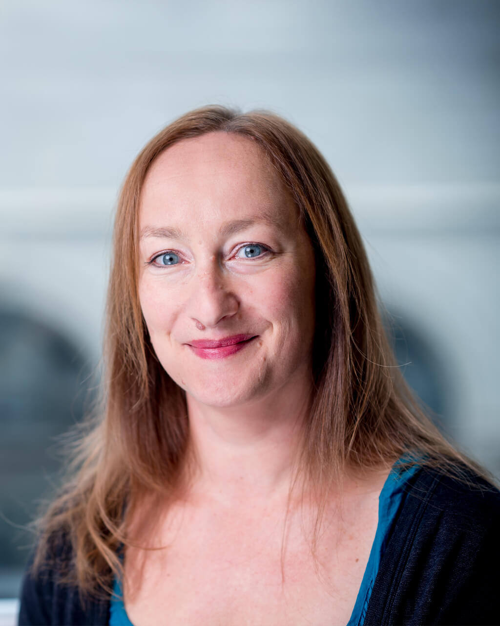 A woman with long light brown hair, wearing a teal top and black sweater, is smiling at the camera with a blurred background.